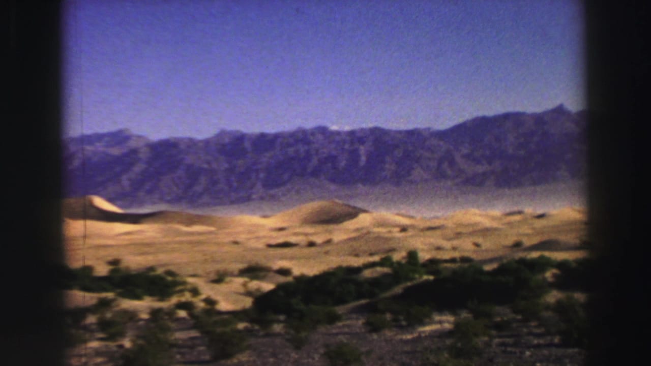 Sand dunes and distant mountains under a blue sky with dark film-frame borders.