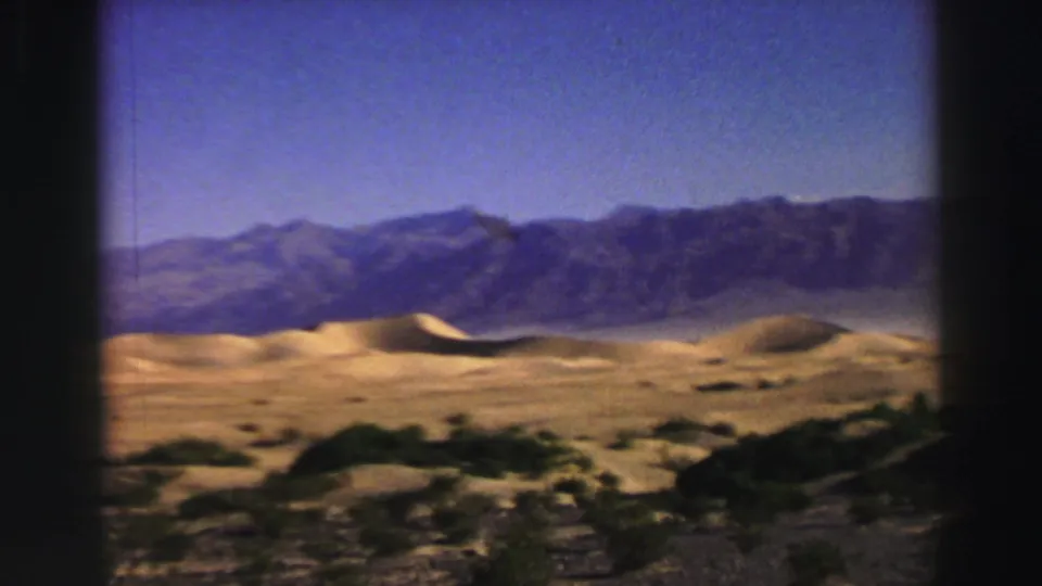 Wide 1969 8mm landscape view with sandy dunes, scattered vegetation, and blue-gray mountains under clear sky.