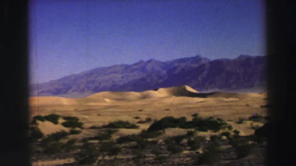 1969 8mm frame with scrubby foreground plants, sunlit dunes, and a rugged mountain ridge in a Western landscape.