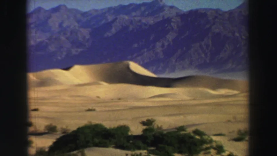 Closer 1969 8mm view of smooth golden dunes, dark low vegetation, and steep blue-gray mountains in the background.