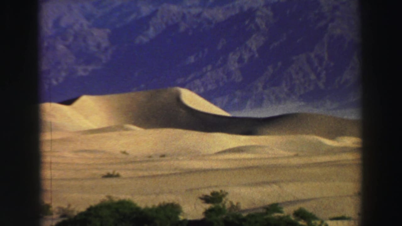 Golden sand dunes and distant mountains under a blue sky, framed by dark 8mm film edges.