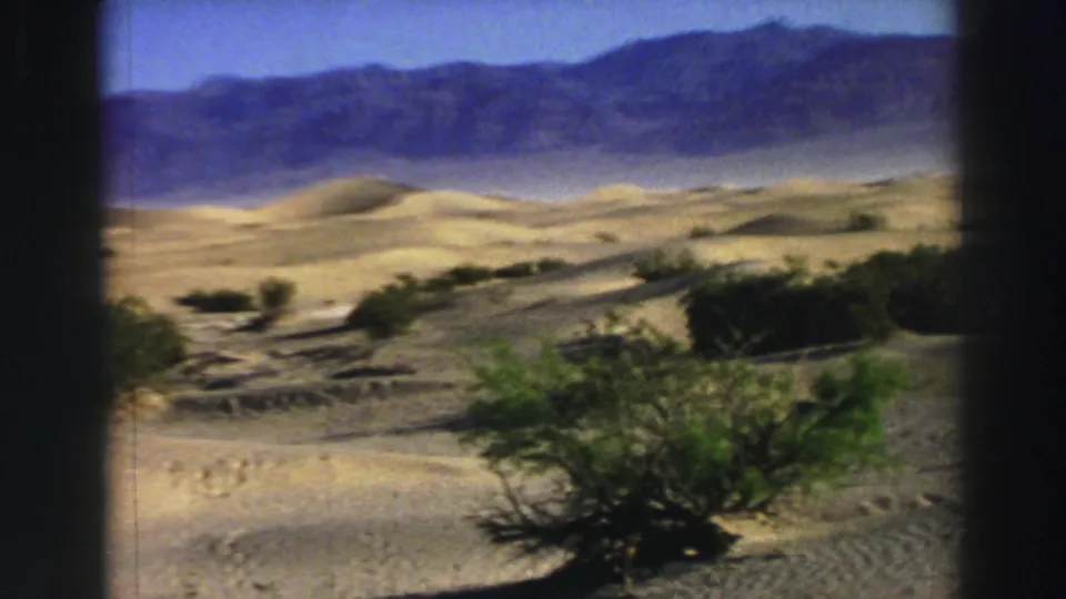 1969 8mm home-movie frame with rippled sand, low shrubs, dunes, and hazy mountains across the horizon.