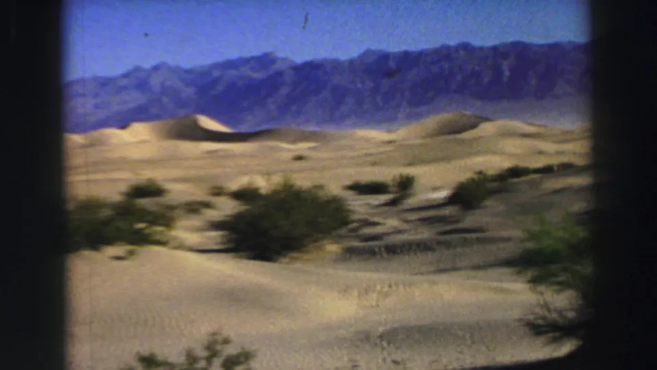 1969 8mm landscape frame showing desert scrub, rolling dunes, and rugged mountains with visible gate-edge darkness.