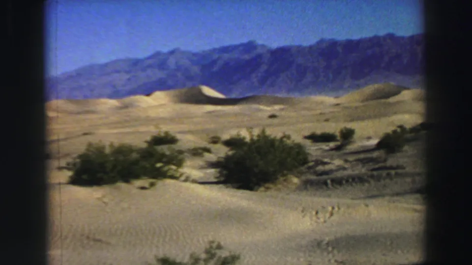 1969 8mm frame from metadata-identified Lake Tahoe, showing tan dunes, low shrubs, and blue-gray mountains in bright daylight.