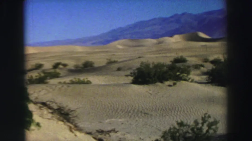 A handheld 8mm view of sandy dunes and scattered desert shrubs, with a distant mountain range beneath clear sky.