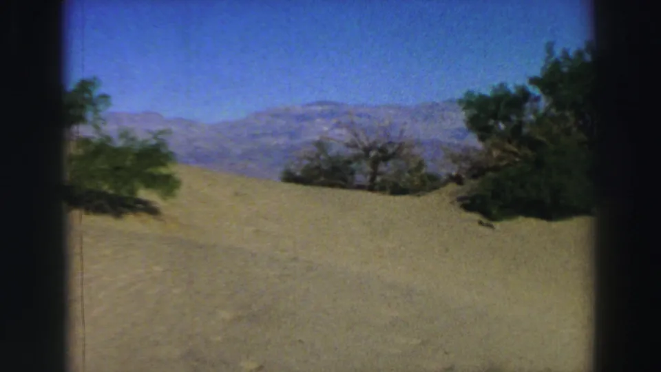 1969 8mm travel footage frame with a sandy rise, sparse trees, and distant mountain ridges near Lake Tahoe.