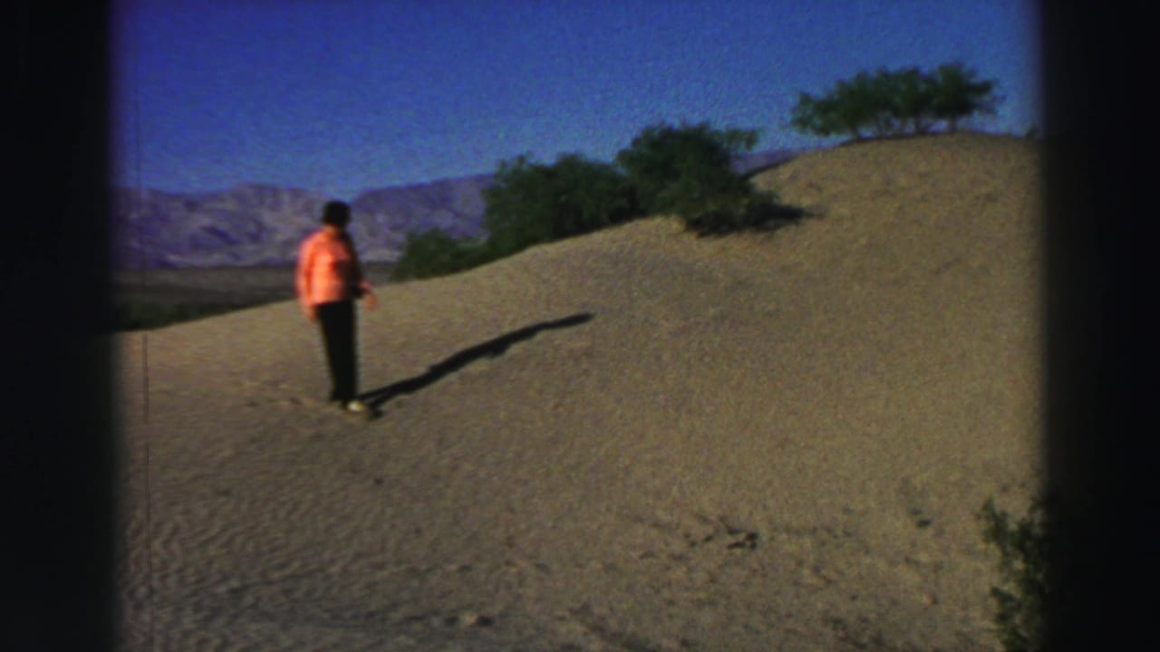 Woman in a peach blouse stands on a sandy hillside with mountains in the distance.
