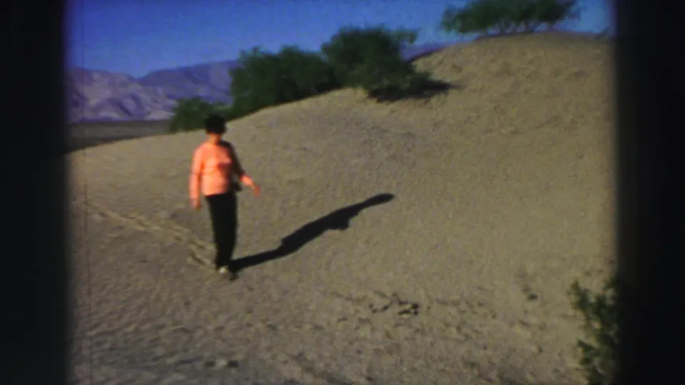 A woman in a peach blouse walks carefully across the sandy hillside in the supplied 1969 Lake Tahoe home-movie scene.