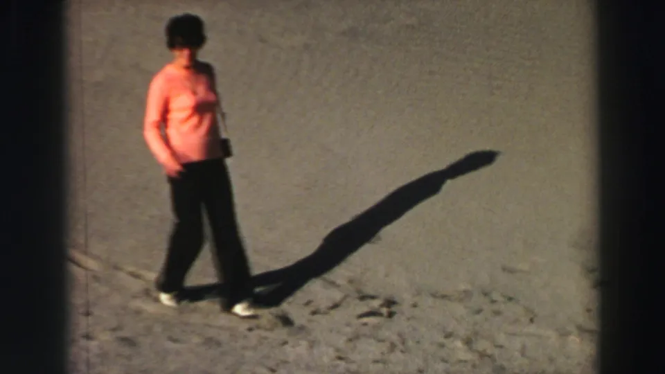 Closer 8mm frame of the woman in a peach blouse walking across sand, her shadow stretched across the Lake Tahoe hillside.
