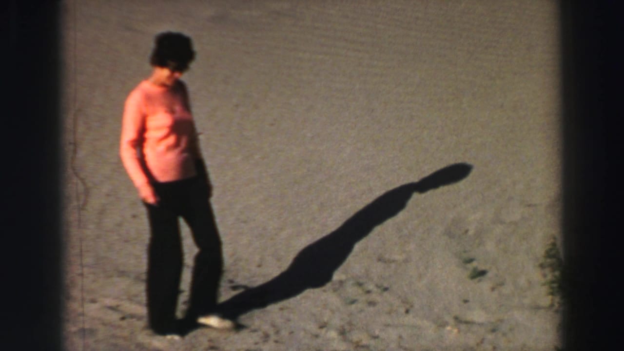 Woman in orange shirt walking on sandy ground with a long shadow.