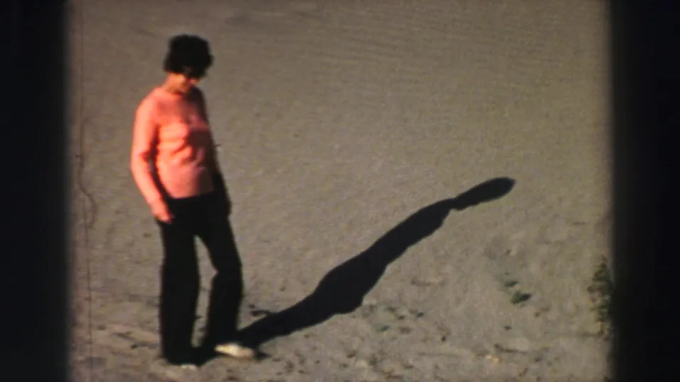 Woman in an orange shirt and dark pants walks across sandy Lake Tahoe terrain in bright 1969 sunlight.