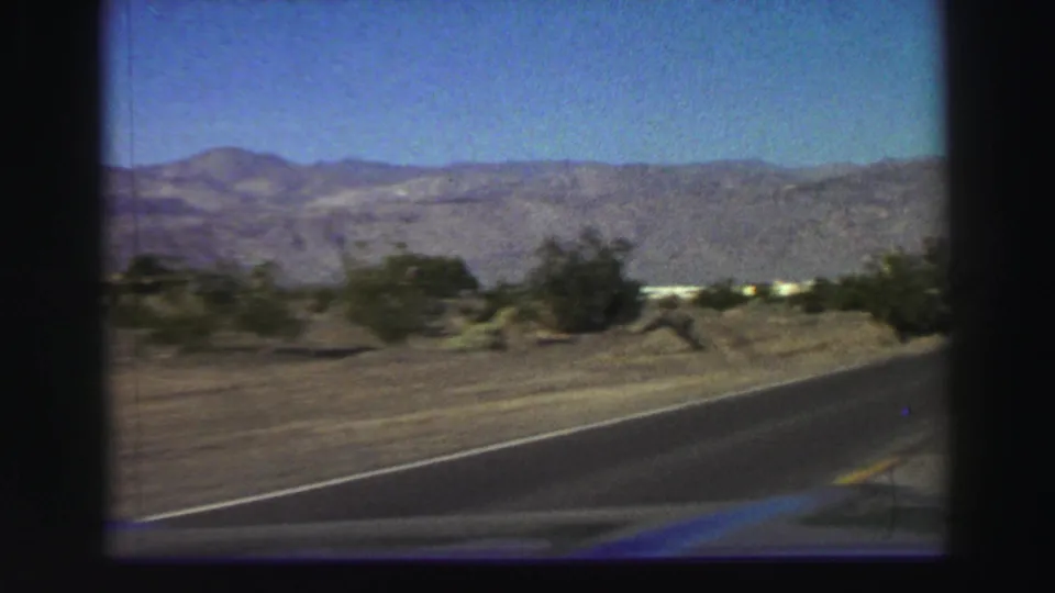 1969 home-movie road shot near Lake Tahoe showing a two-lane paved road, low shrubs, dry ground, and mountain ridges.
