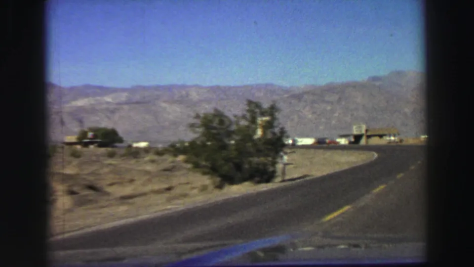 Late-1960s 8mm vehicle view near Lake Tahoe with a curving road, roadside shrubs, low buildings, and mountain backdrop.