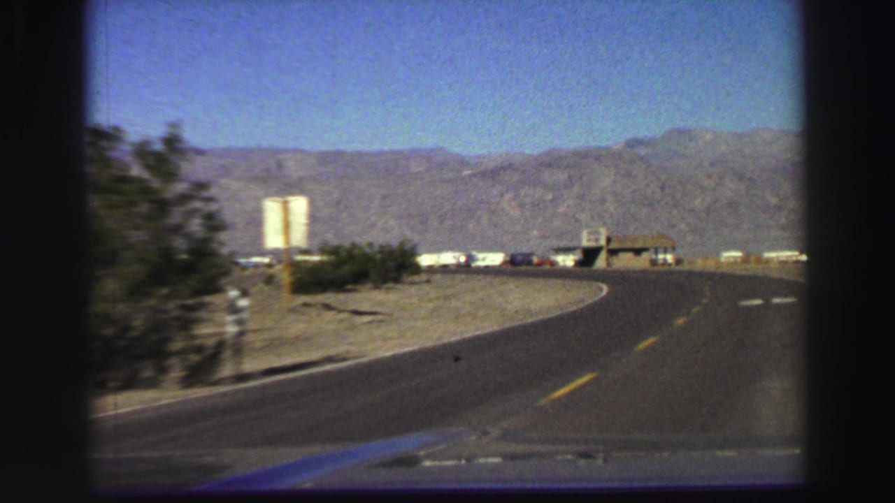 Two-lane road curving past shrubs toward distant mountains and roadside structures.