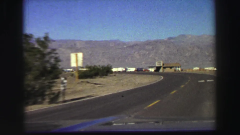 1969 Lake Tahoe-area road view from a moving vehicle, with a curving two-lane road, yellow center line, shrubs, signs, buildings, and mountains.