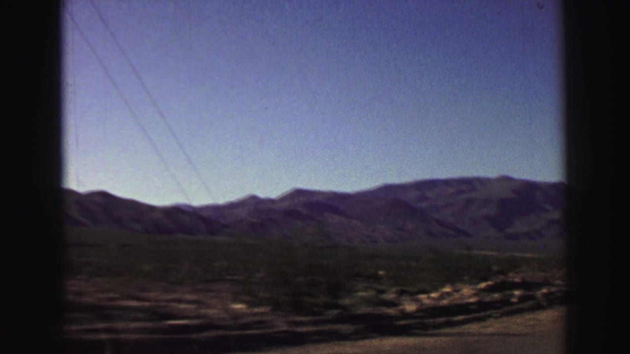 Mountain range seen through a car window beside a paved road and dry roadside terrain.