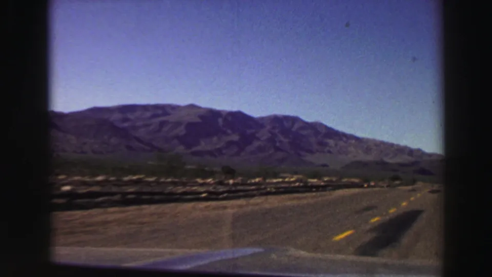 1969 8mm road-trip frame looking ahead from a car toward a paved highway, arid foreground, and mountain range near Lake Tahoe.