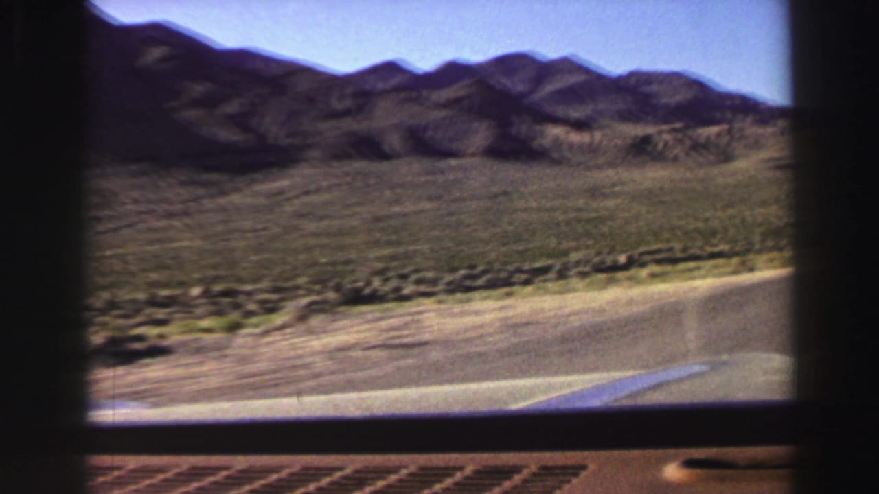 1969 Lake Tahoe-area home-movie frame showing a forward car view along a paved road bordered by scrubland and low mountain ridges.