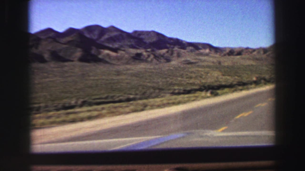 Curving paved road with yellow center lines, shrubs, and mountains beyond.