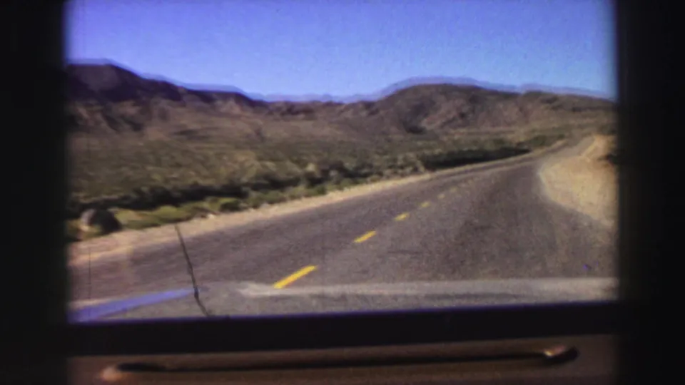 Home-movie driving view of a two-lane road bending through dry California mountain terrain, with sparse roadside vegetation and distant hills.