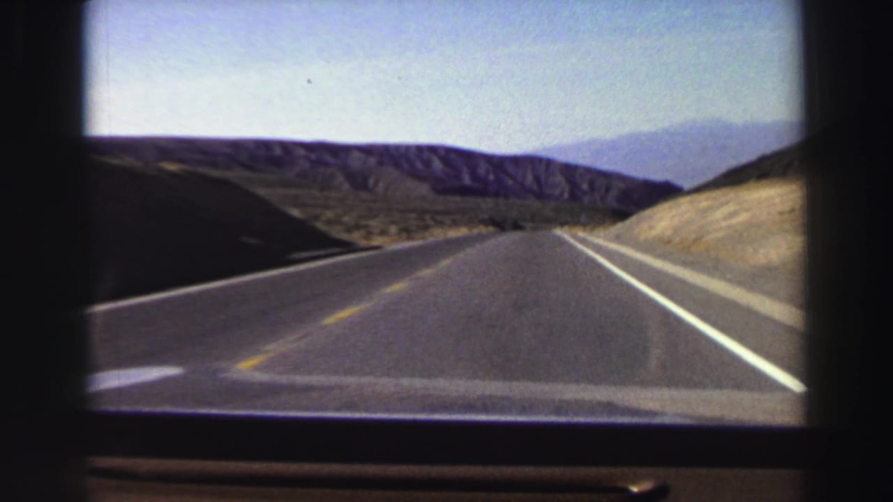 1969 8mm home-movie view from a car on a two-lane road near Lake Tahoe, with arid hills and distant mountains ahead.