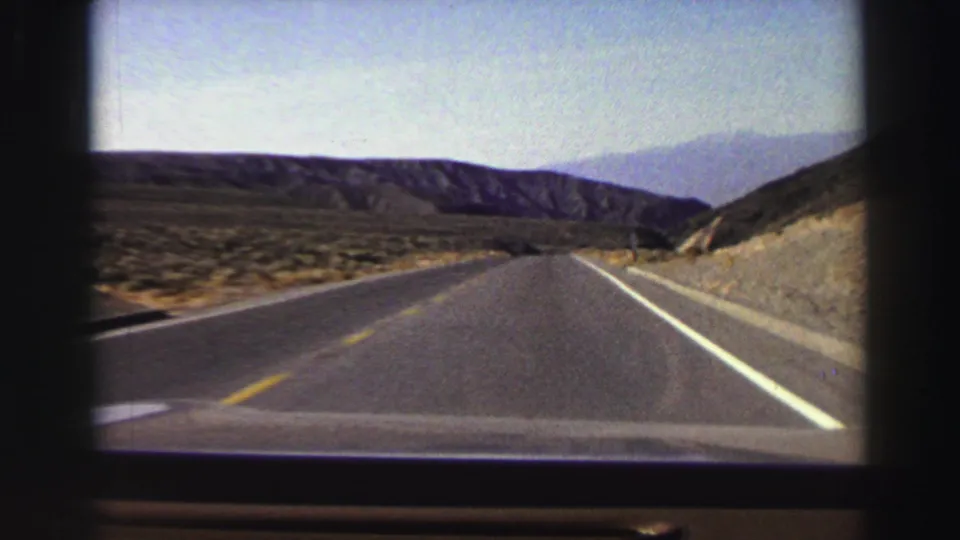 First-person 8mm view of a road curving through dry Lake Tahoe-area hills, with mountains in the distance and no people visible.