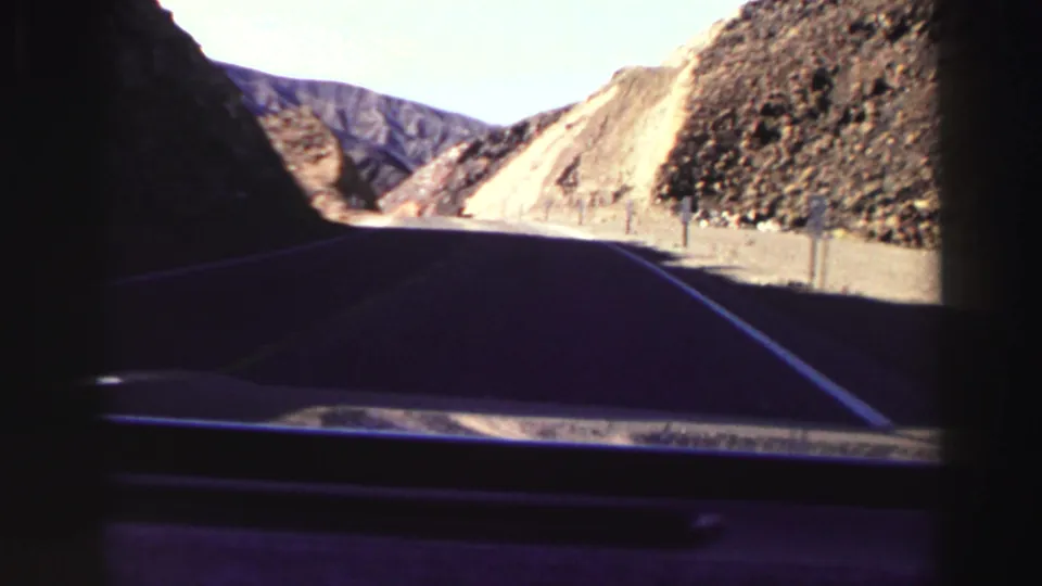1969 road-trip POV from inside a car entering a shaded curve between rocky, sunlit hills in the Lake Tahoe area.