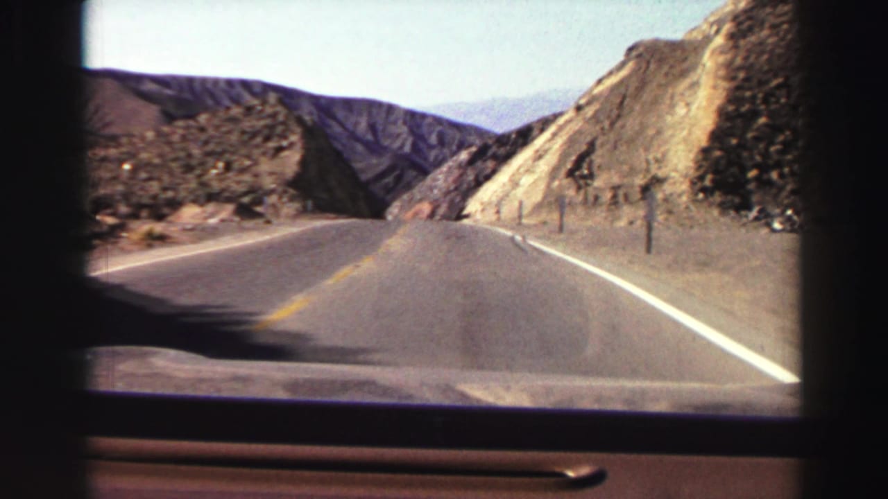 Forward view through a car windshield on a two-lane road between rocky mountain slopes.