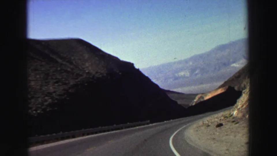 1969 Lake Tahoe-area road-trip view from a moving vehicle, with a paved curve between rocky slopes and distant mountains.