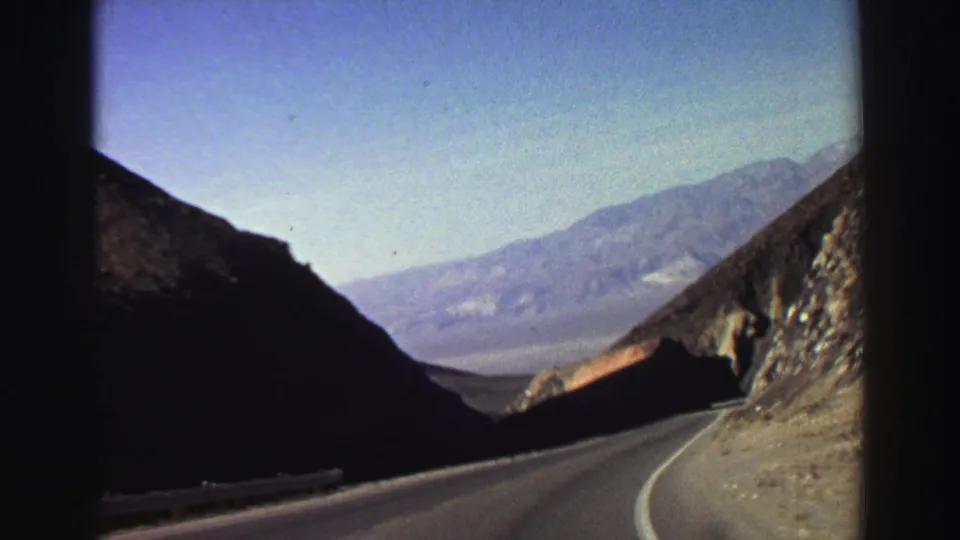1969 Lake Tahoe-area home-movie frame showing a curving paved road, shadowed rock walls, and hazy distant mountains.