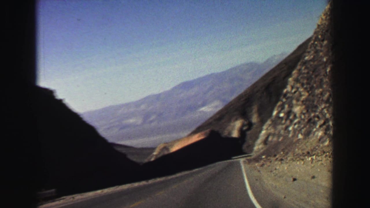 Two-lane mountain road curves between rocky slopes with distant ridges under blue sky.