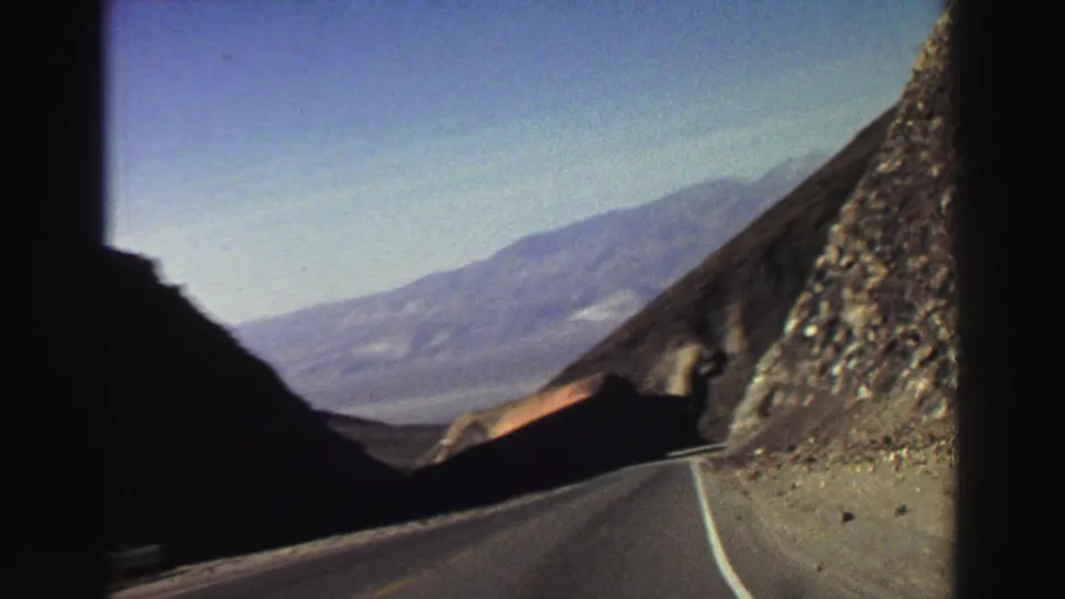 1969 8mm Lake Tahoe-area road view from a moving car, showing a paved highway between steep rocky mountains and hazy distant ridges.