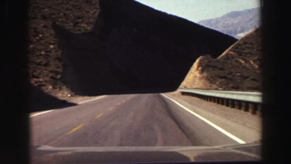 1969 home-movie frame of a paved mountain road entering a darker canyon-like section, with a guardrail and rocky slopes visible.