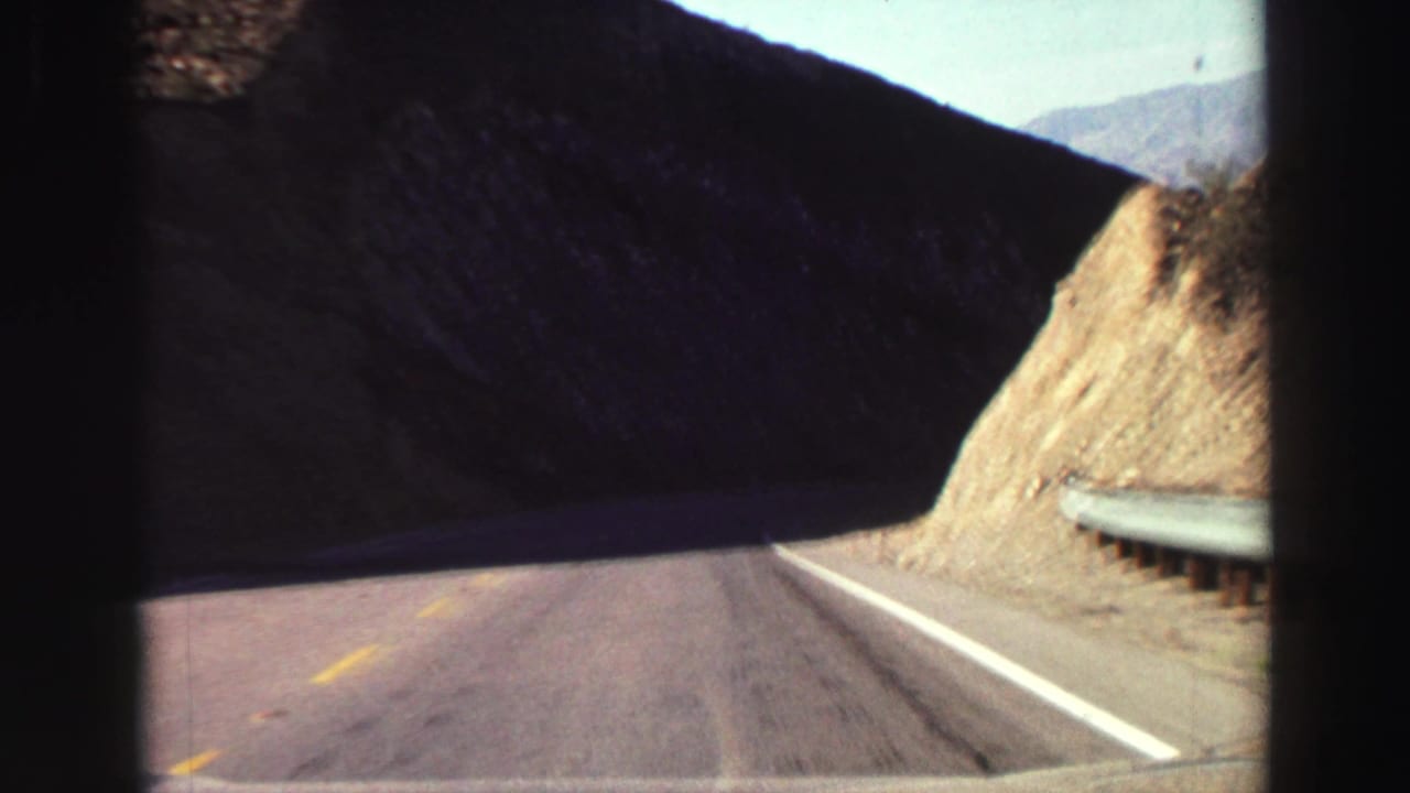 Two-lane road curves past a rocky hillside with a guardrail at the shoulder.
