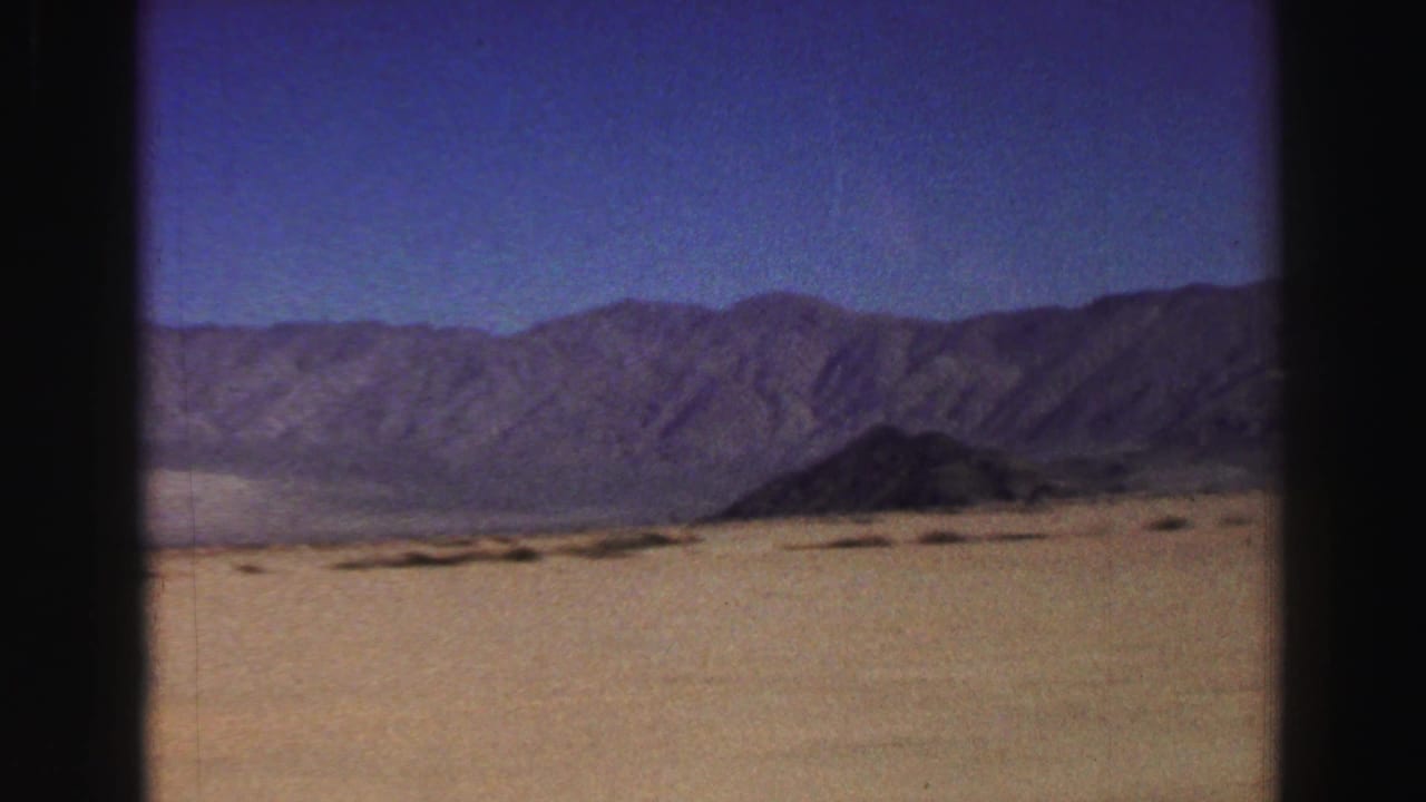 Desert plain stretches toward a mountain range under a clear blue sky.