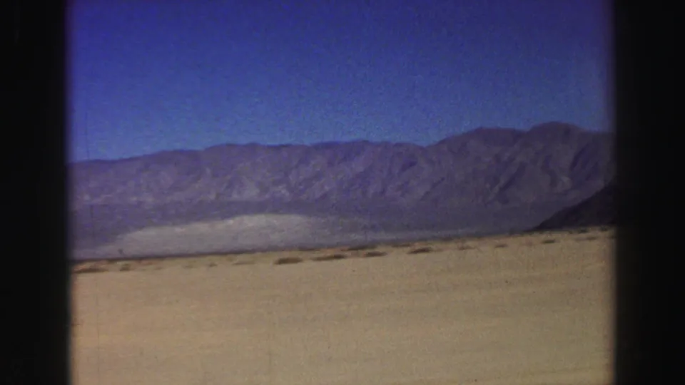 1969 8mm travel footage frame with open arid ground, sparse vegetation, and a broad mountain range viewed from a moving car in California.