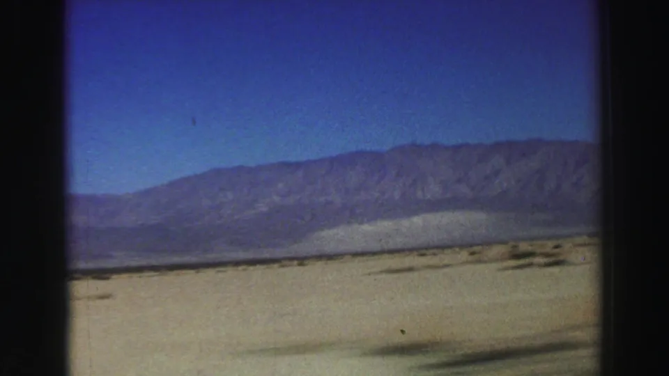 1969 8mm home-movie view of a pale arid basin and distant mountain ridge, with dark interior window borders indicating the shot was taken from inside a vehicle.