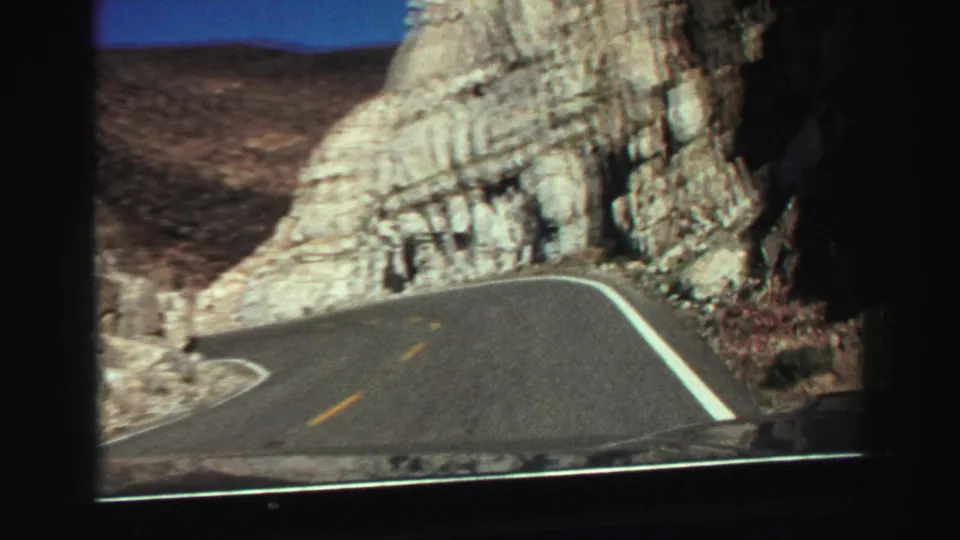 Vintage 8mm driving view of a left-curving road through rugged rock cuts in the Lake Tahoe, California area.
