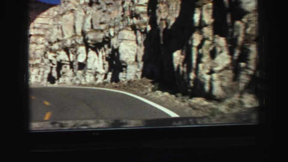 1969 8mm road footage showing a paved mountain road beside a tall rock wall, filmed from inside a moving vehicle.