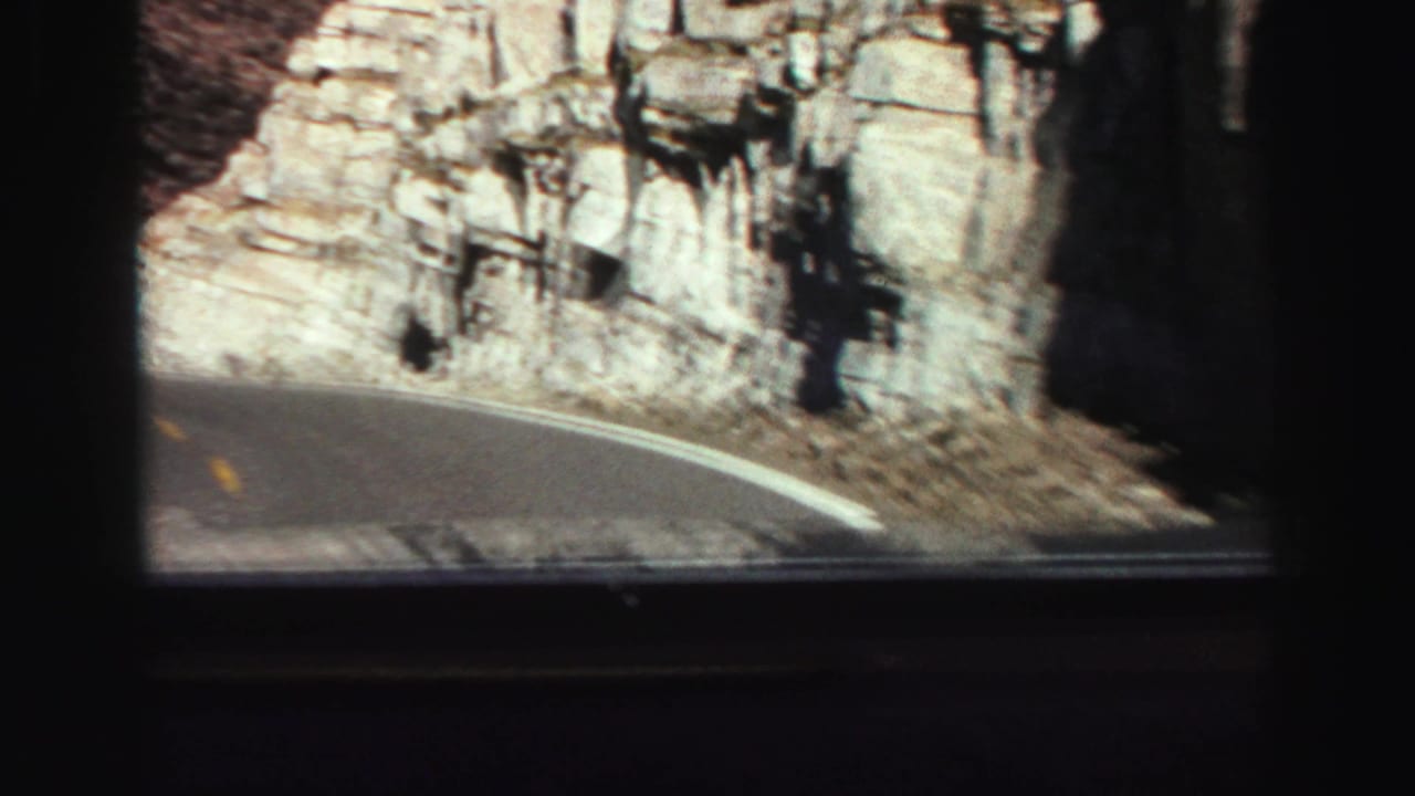 Curving mountain road seen through a vehicle window beside pale rocky cliffs.