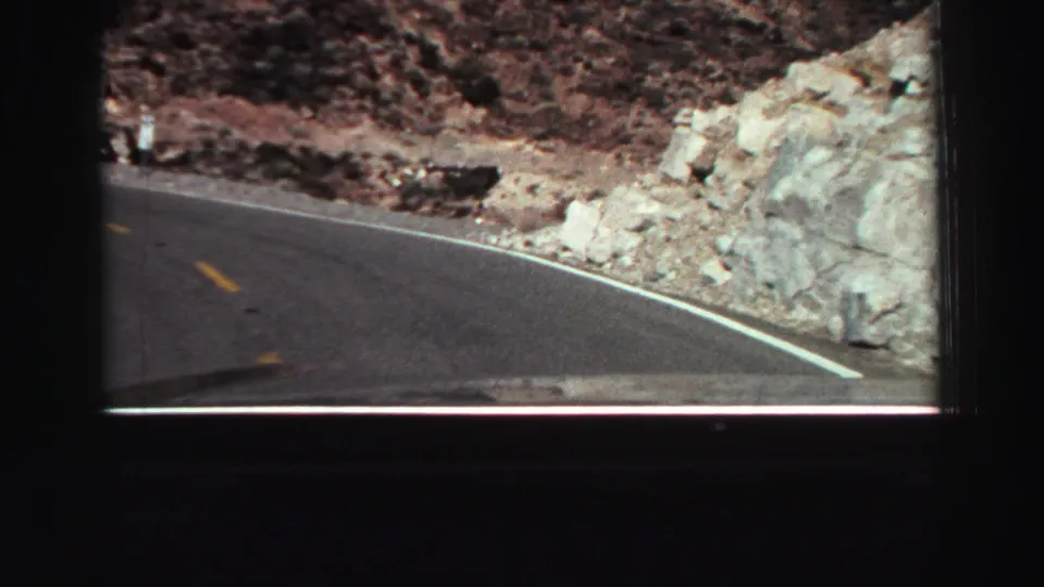Passenger-view 8mm frame of a Lake Tahoe-area mountain road, with painted lines, scattered boulders, and dry rocky slopes beside the pavement.
