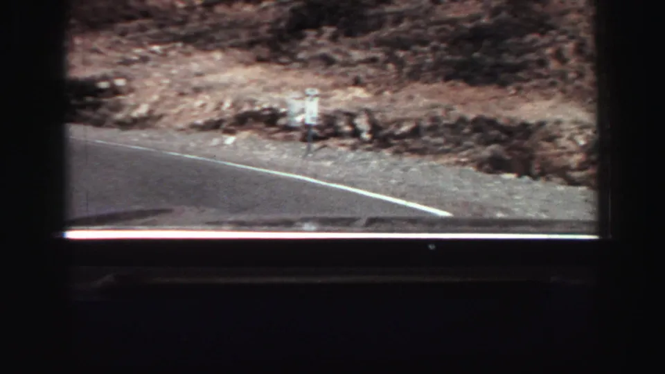A 1969 Lake Tahoe-area road-trip frame shows the vehicle passing a curving road edge with rocky shoulder and dry hillside beyond.