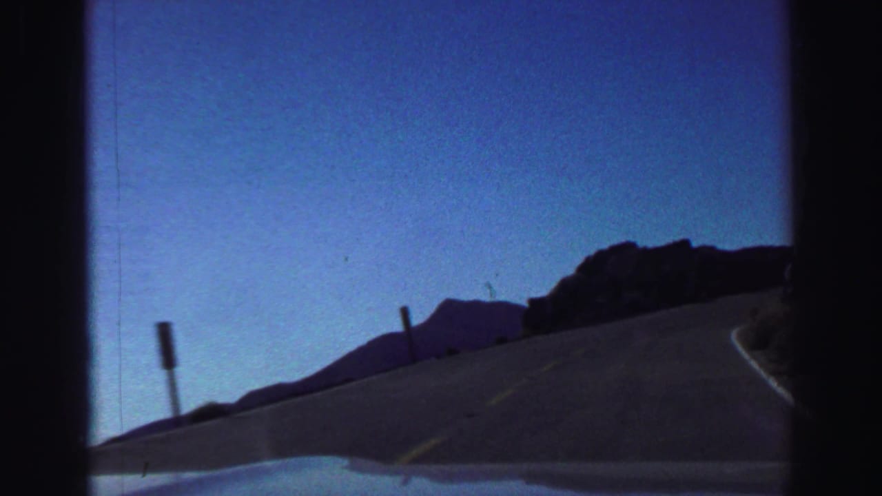 Two-lane mountain road curving past rocky slopes under a blue sky.