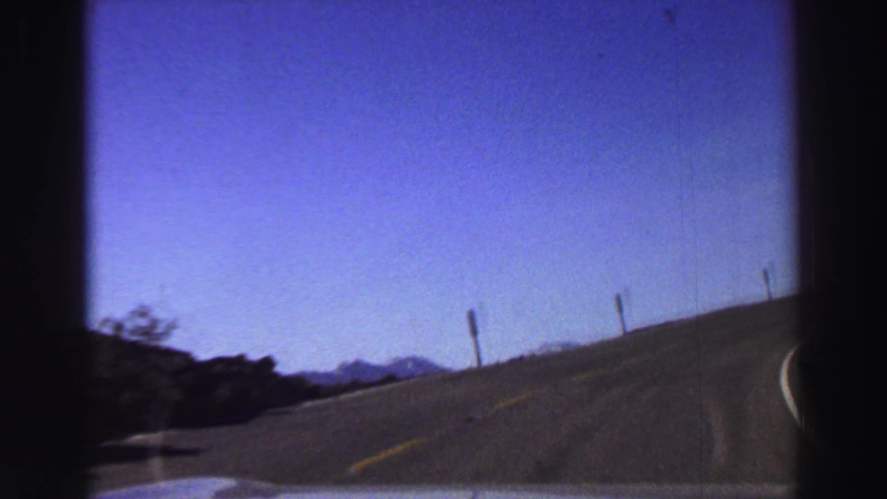 Curving two-lane mountain road seen from a moving car under blue sky.