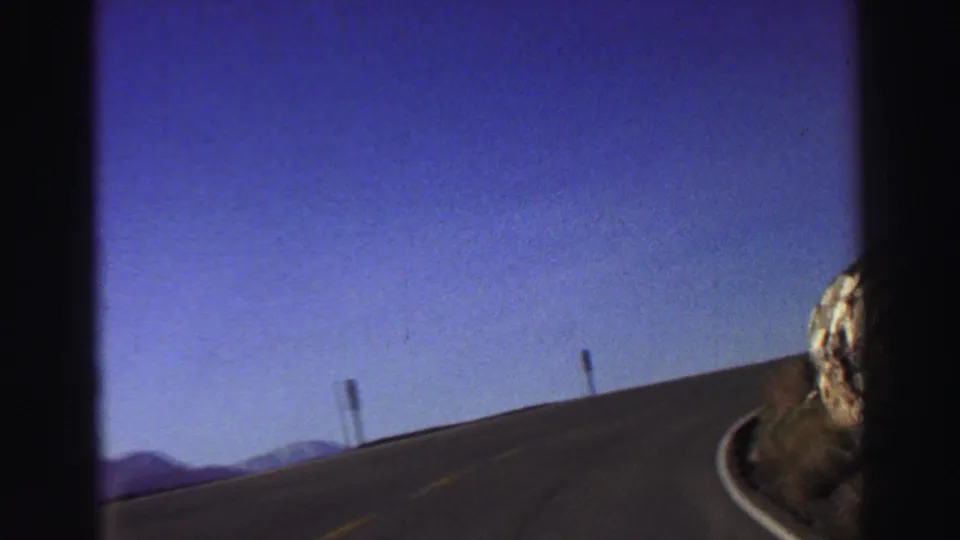 Passenger-view 8mm frame of a Lake Tahoe mountain road curving past roadside posts and a rocky slope in 1969 footage.