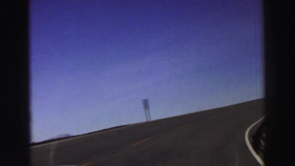 1969 Lake Tahoe home-movie frame showing a paved mountain road, a roadside sign, and open sky from a moving car.