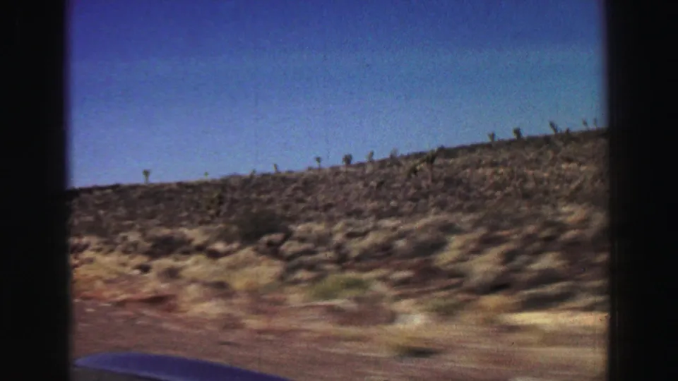 Moving 8mm roadside view near Lake Tahoe with scrub-covered hillside, blue sky, and a small car hood edge.