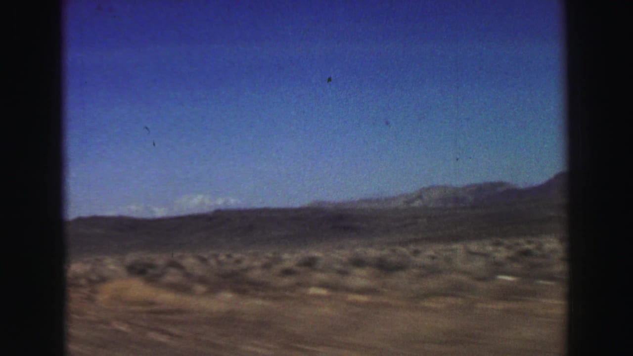 1969 8mm road-trip frame showing arid roadside scrub, distant mountains, and blue sky near the Lake Tahoe, California metadata location.
