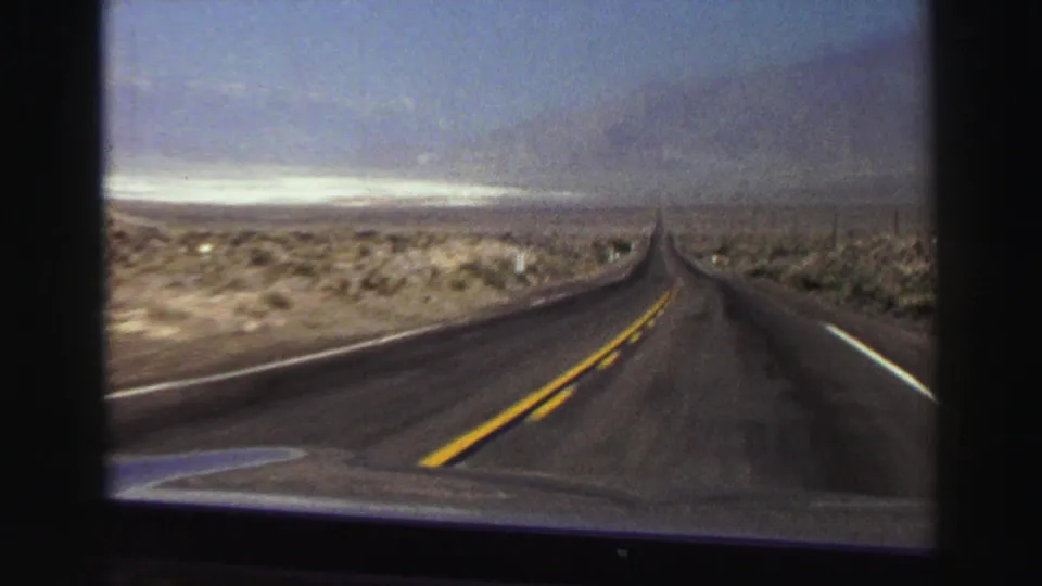Forward-facing 1969 8mm view of a quiet two-lane highway with yellow center lines, white road edges, desert scrub, and distant mountains.