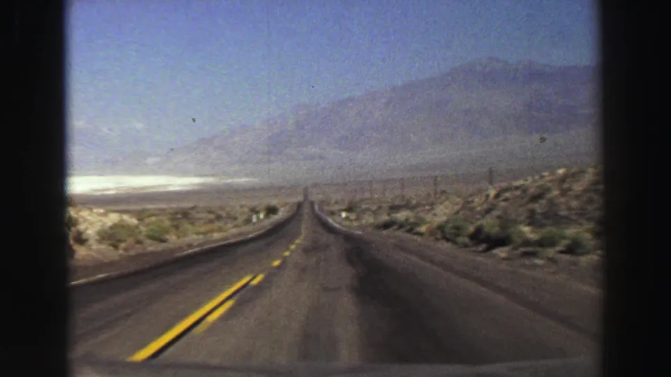 1969 8mm frame of a straight road through dry terrain, with yellow dividing lines, sparse shrubs, and hazy mountains in the distance.